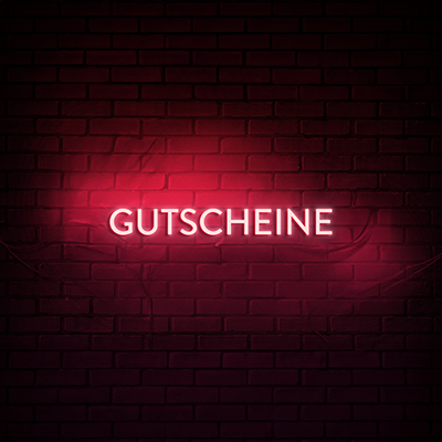 Das Bild zeigt den Schriftzug 'GUTSCHEINE' in leuchtenden Neonbuchstaben, die an einer Ziegelmauer befestigt sind. Der Text erstrahlt in einem roten Neonlicht.