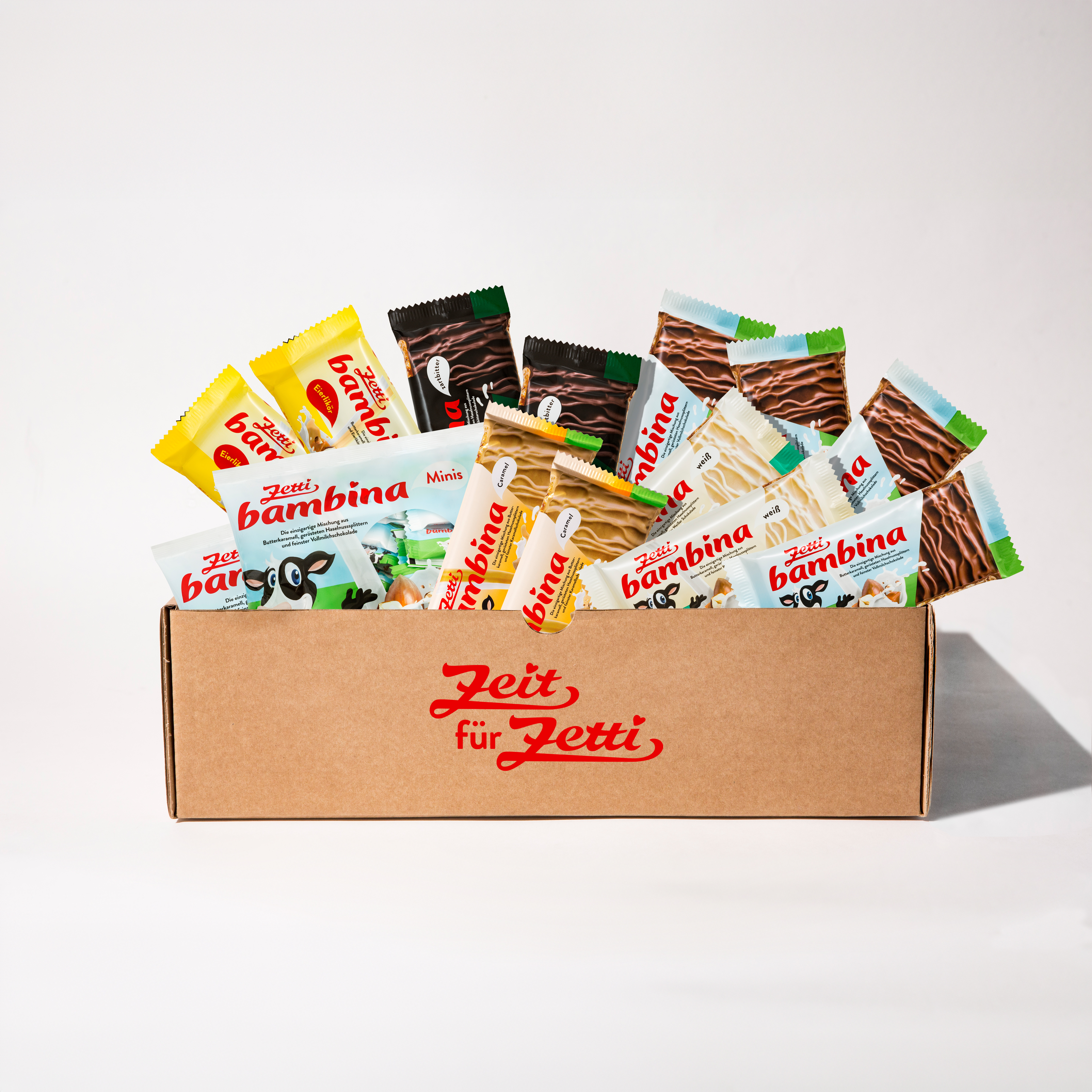 Zetti Bambina Power Das Bild zeigt einen Karton mit dem Schriftzug 'Zeit für Zetti' in roter Schrift. Der Karton ist gefüllt mit verschiedenen Zetti Bambina Riegeln in verschiedenen Sorten, darunter weiße, dunkle und milchschokoladene Varianten, sowie Zetti Bambina Minis