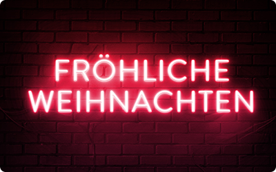 Eine Gutscheinkarte zeigt den Schriftzug 'Fröhliche Weihnachten' in leuchtend roten Neon-Großbuchstaben vor einem schwarzen Ziegelmauer-Hintergrund.
